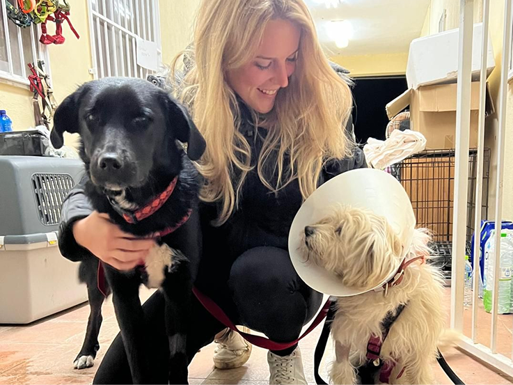 Lara Poppe im Tierschutz-Engagement: Sie lächelt liebevoll zwei Hunde an, die sie umarmt, während der kleinere eine Halskrause trägt.